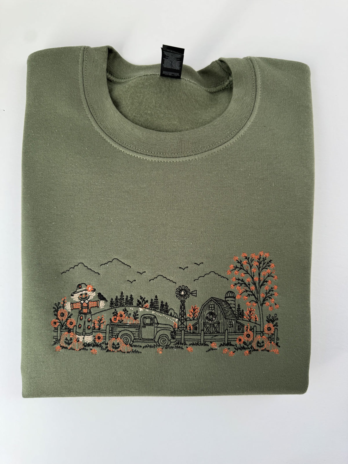 Autumn Farm line art embroidered on a Military Green crewneck: scarecrow, pickup truck, windmill, barn and orange pumpkins