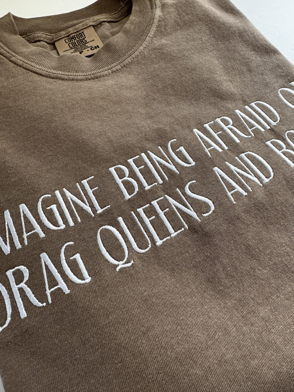 "Imagine being afraid of drag queens and books" embroidery shown at an angle on an Espresso tee