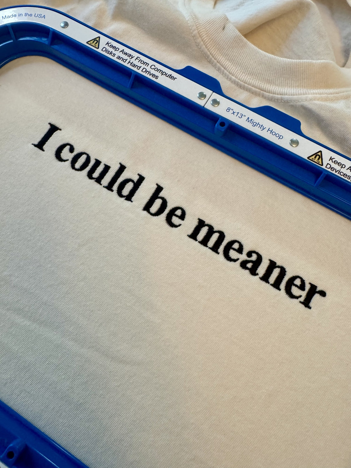 The "I could be meaner" design stitched on an Ivory tee, still clamped in the embroidery hoop