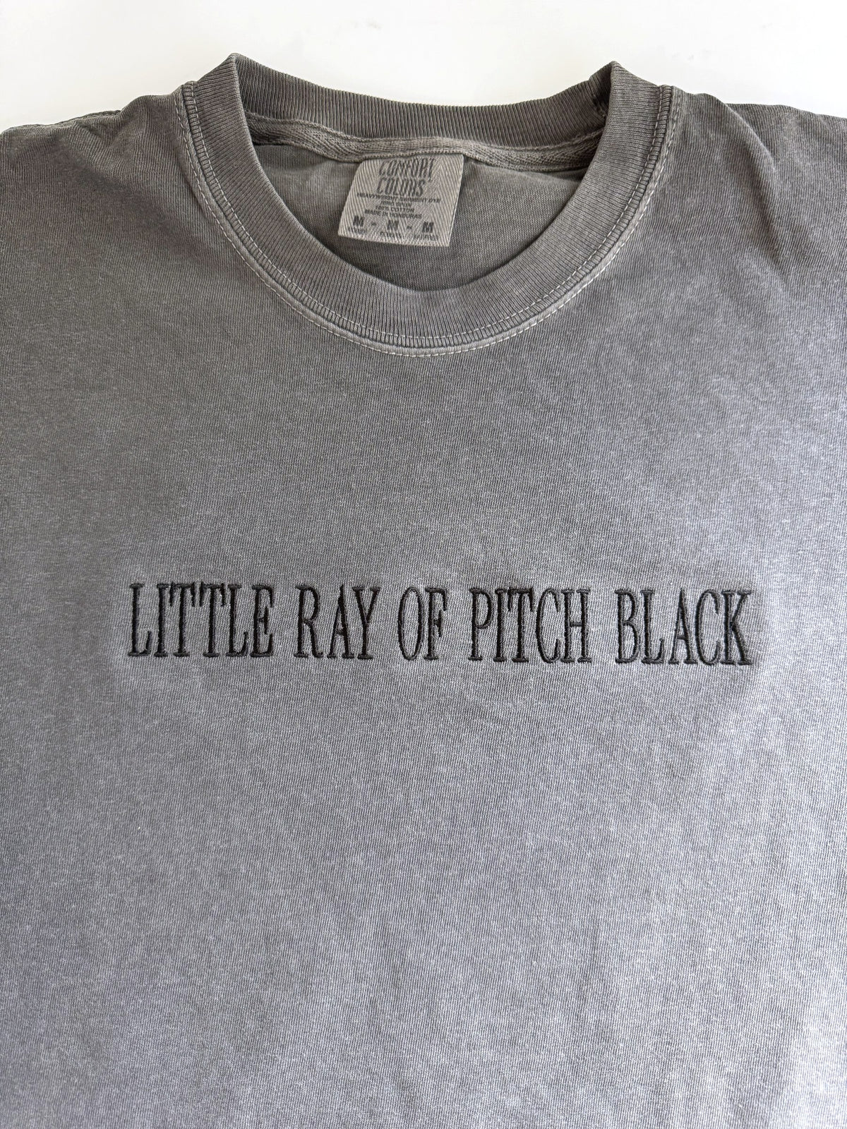 "Little ray of pitch black" embroidered tee in Pepper, laid flat, stitched tonal black on grey