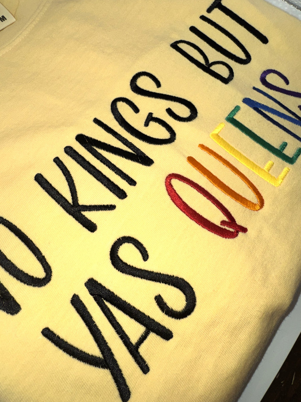 "No kings but yas queens" embroidery shown at an angle, black lettering with rainbow thread
