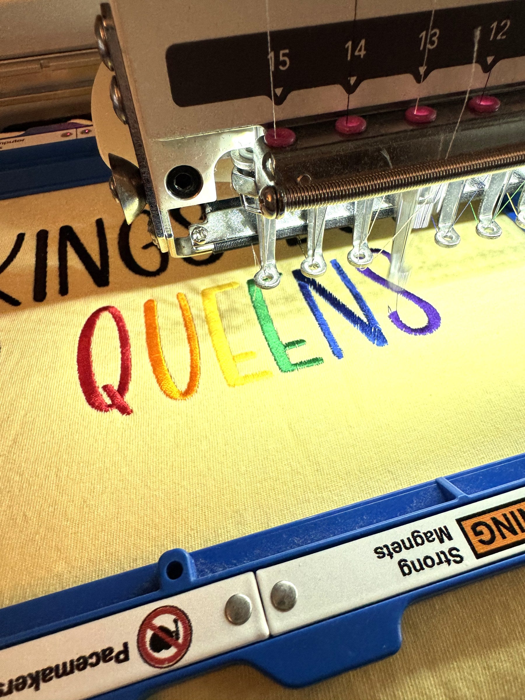The "No kings but yas queens" tee on the embroidery machine, rainbow lettering part-stitched