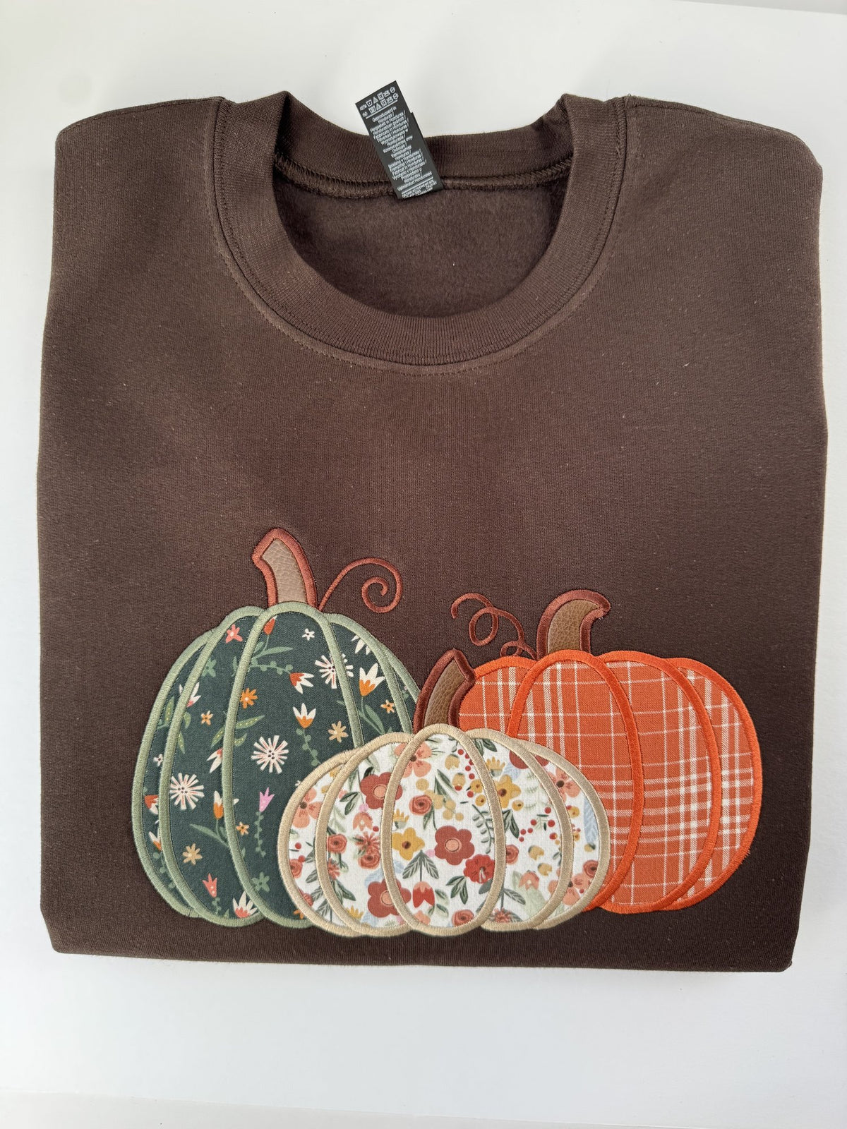Pumpkin Trio appliqué on a Dark Chocolate crewneck: three pumpkins in green floral, cream floral and orange plaid fabric