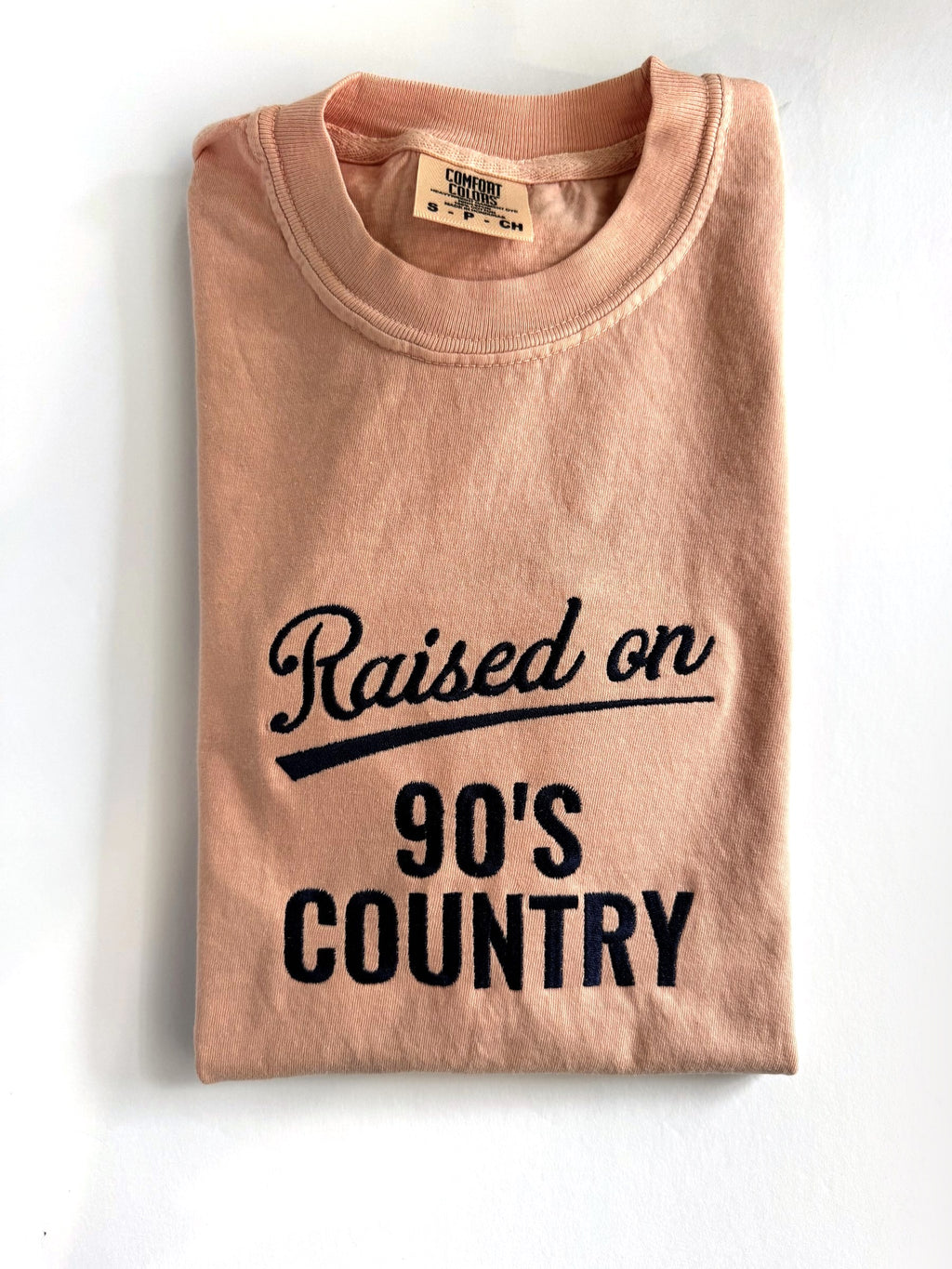 "Raised on 90's Country" embroidered tee in Peachy, folded flat with the collar showing