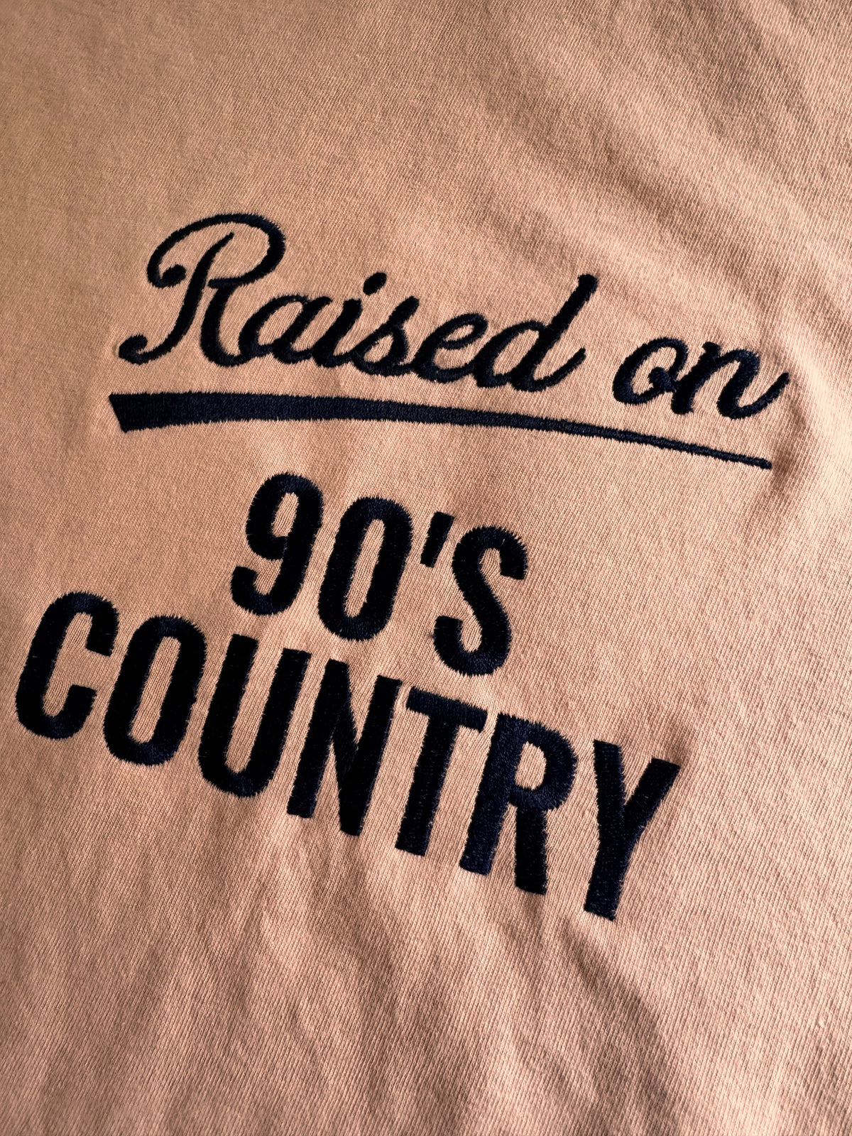 Close-up of the navy embroidered lettering on the "Raised on 90's Country" tee in Peachy