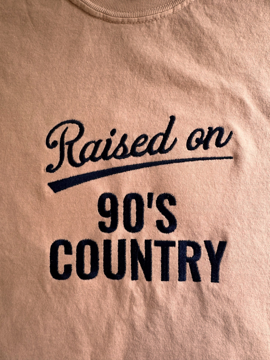"Raised on 90's Country" script and block embroidery on a Peachy tee, shown close