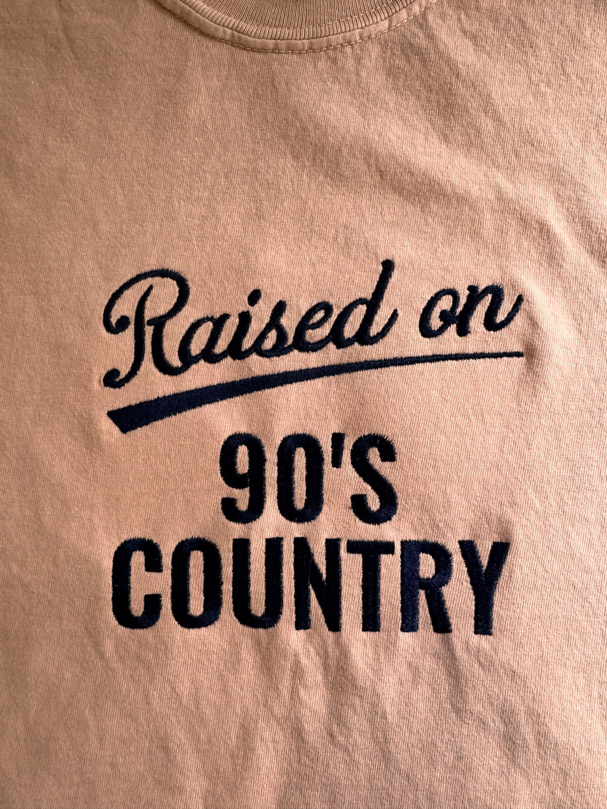 "Raised on 90's Country" script and block embroidery on a Peachy tee, shown close