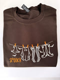 Spooky clothesline embroidered on a Dark Chocolate crewneck: witch hat, ghost, fishnets and boots on a line with orange clothespins