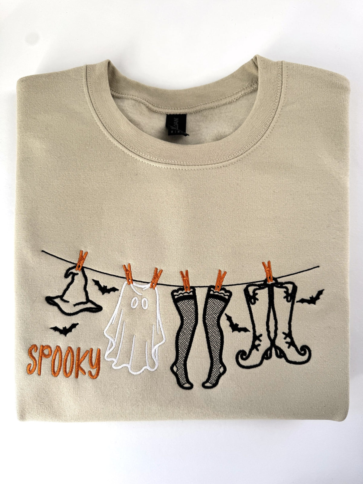 Spooky clothesline embroidered on a Sand crewneck in black outline with a white ghost and orange clothespins