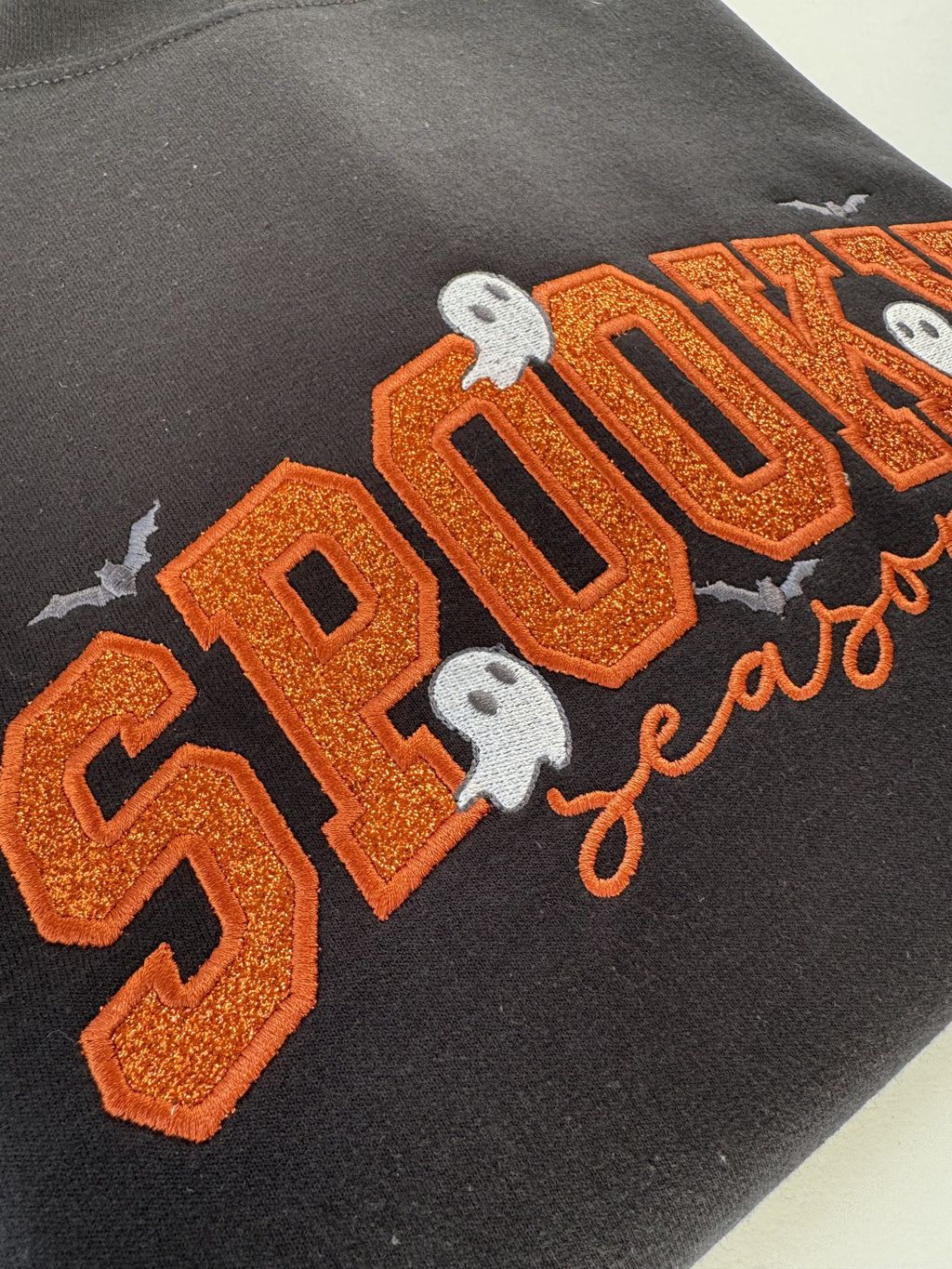 Close up of the orange glitter appliqué letters and stitched ghosts on the Black Spooky Season crewneck