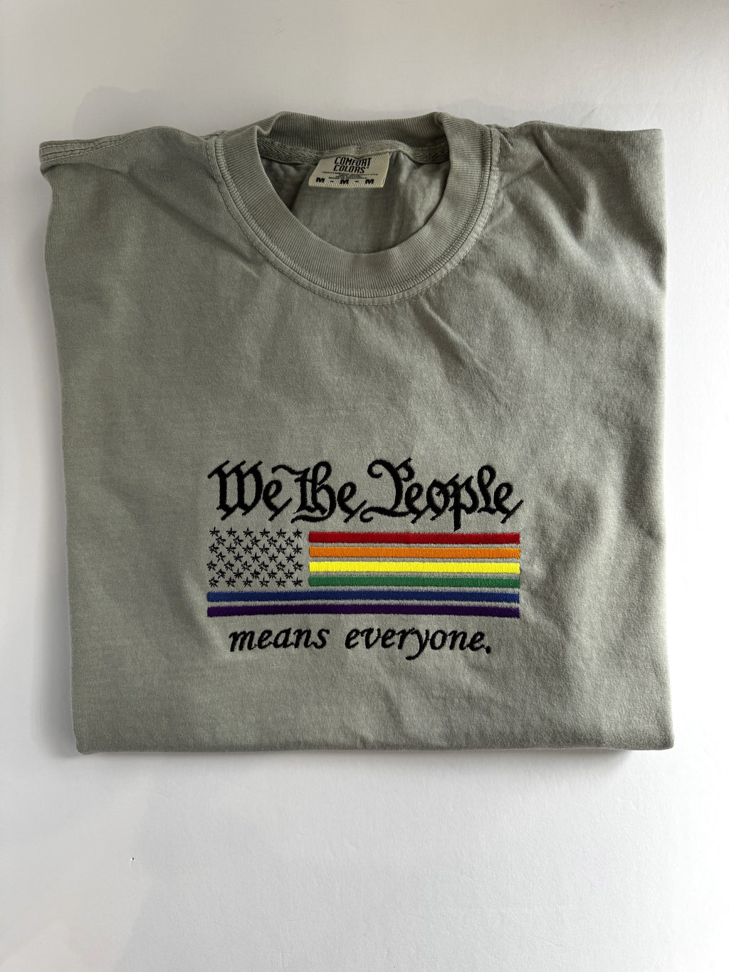 "We The People means everyone" embroidered tee, folded flat with the collar showing, rainbow flag across the chest