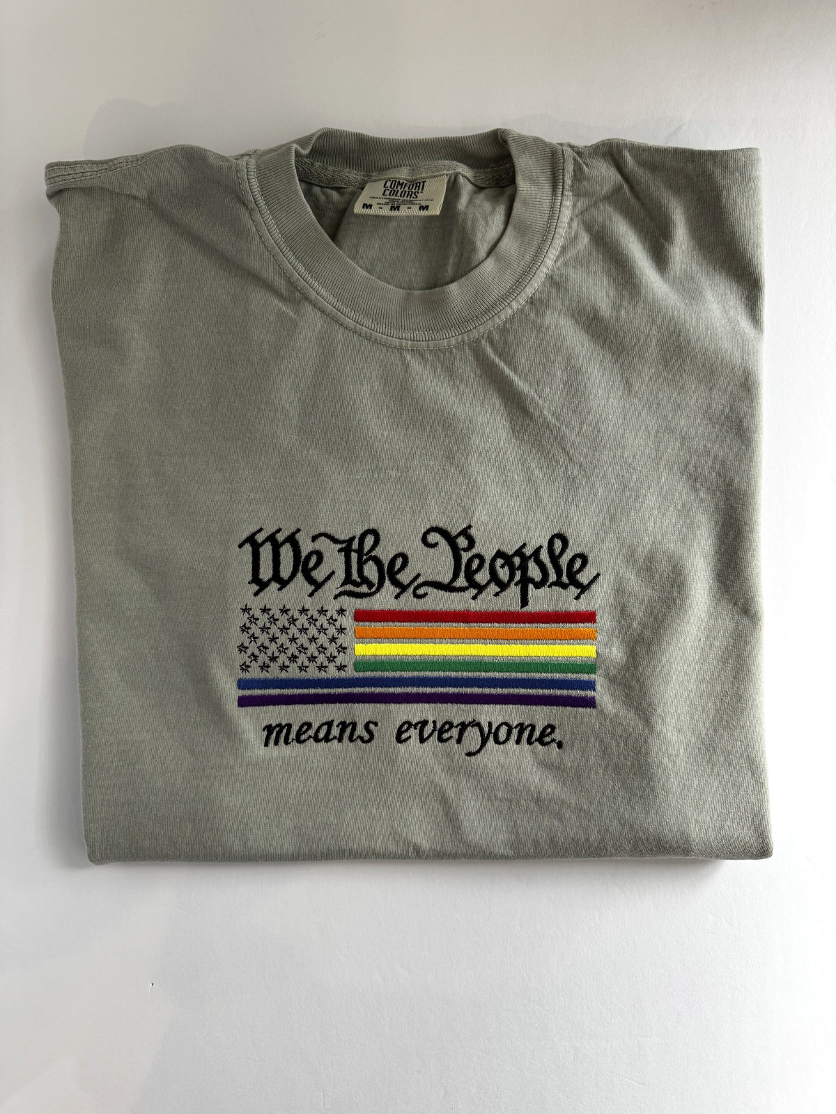 "We The People means everyone" embroidered tee, folded flat with the collar showing, rainbow flag across the chest