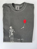 Skeleton watching a red balloon float away with Well, shit embroidered in white on a Pepper tee