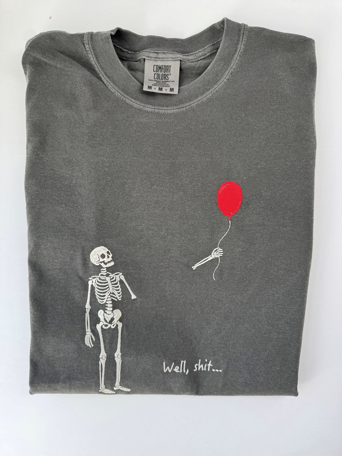 Skeleton watching a red balloon float away with Well, shit embroidered in white on a Pepper tee