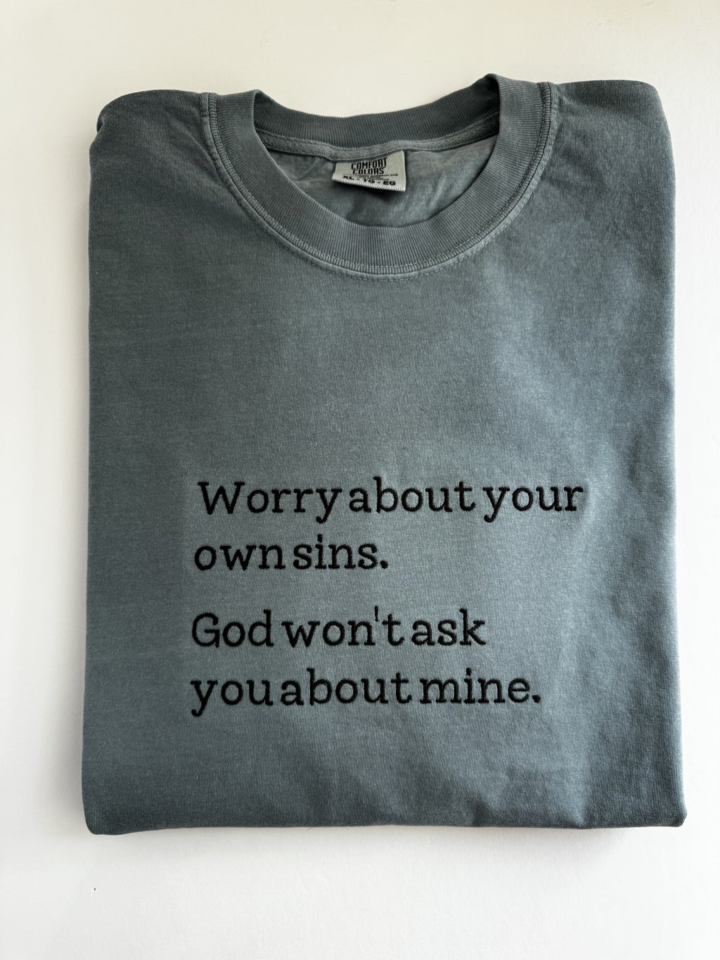 "Worry about your own sins. God won't ask you about mine." embroidered tee, folded flat with the collar showing