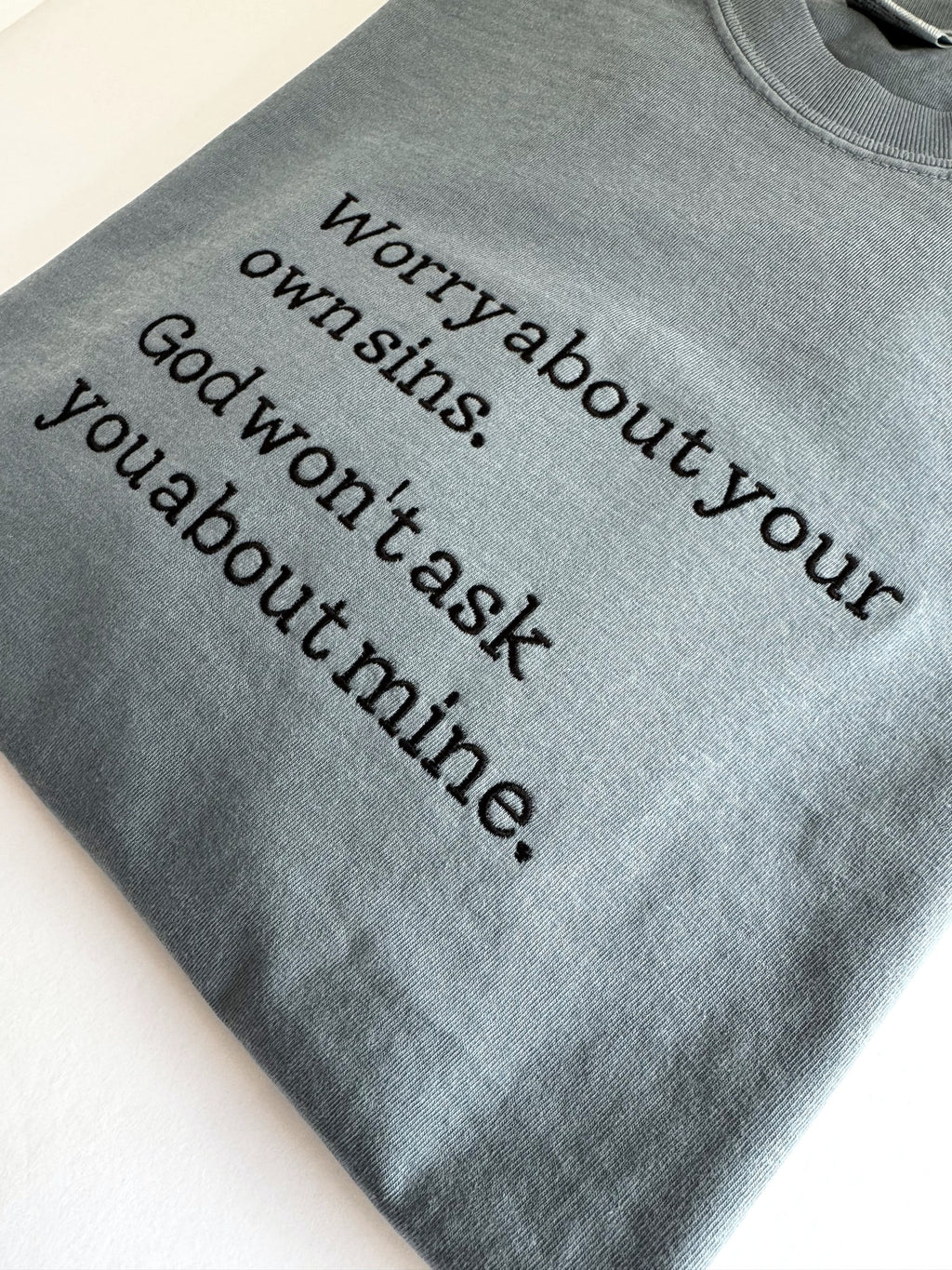 "Worry about your own sins" embroidered tee shown at an angle, dark typewriter lettering